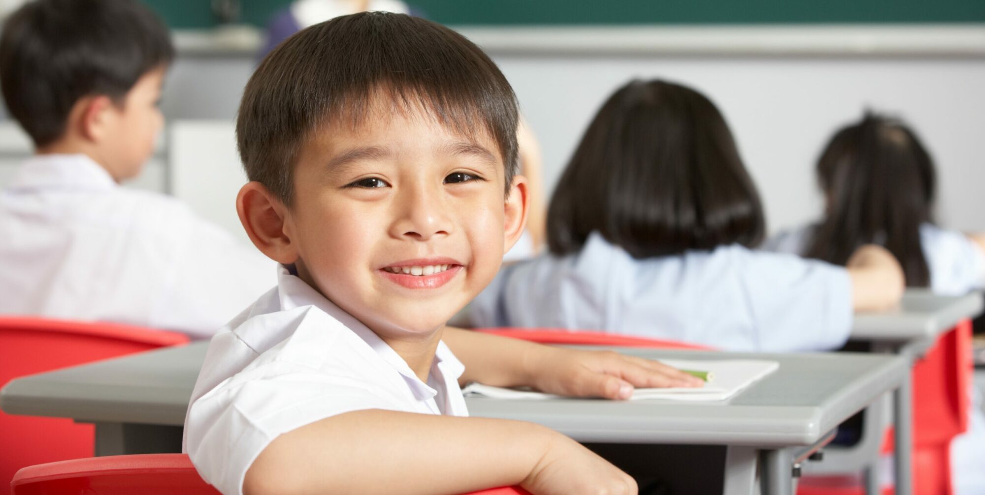 Young smiling child in classroom sitting at a desk with other students.